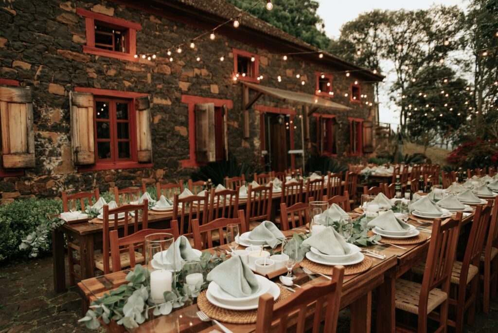 Rustic outdoor wedding reception with long wooden tables and string lights beside a stone farmhouse