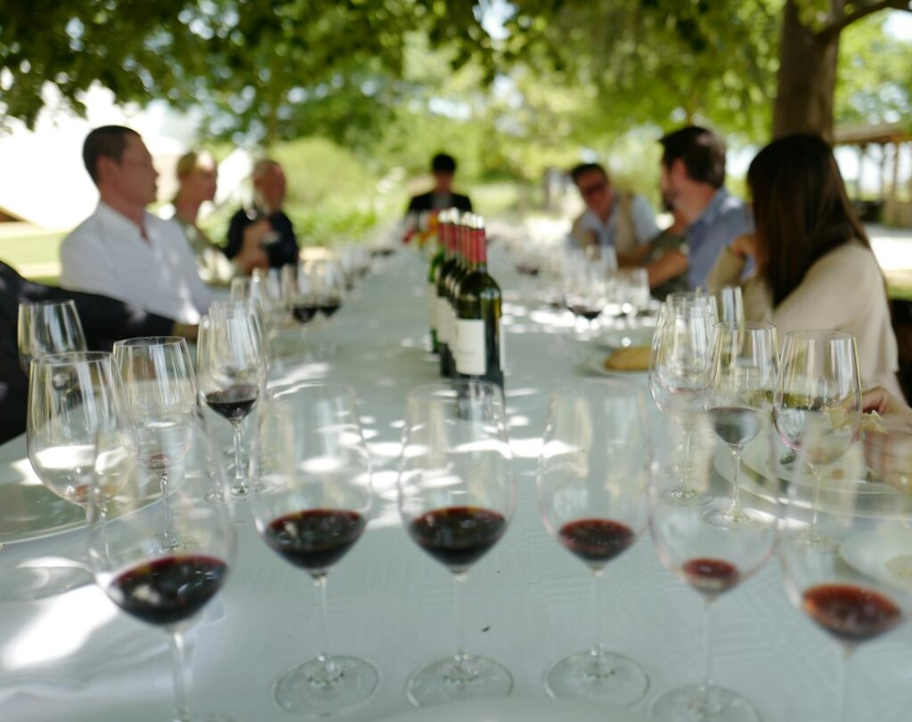 Outdoor wine tasting at a long white-linen table under trees at a Bordeaux vineyard, with rows of wine glasses filled with red and white wine
