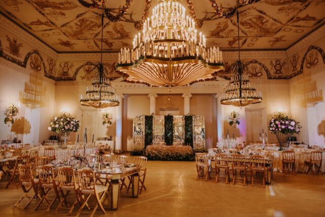 Grand luxury wedding ballroom with crystal chandeliers and warm golden-pink light illuminating elegant reception tables