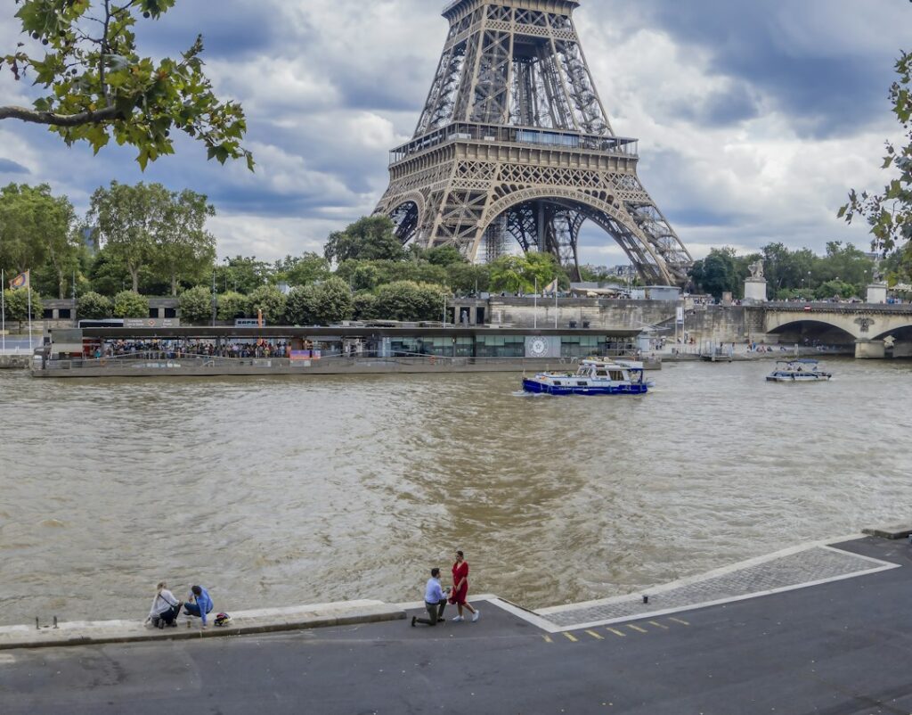 A person kneeling to propose to their partner by the Seine river with the Eiffel Tower behind them